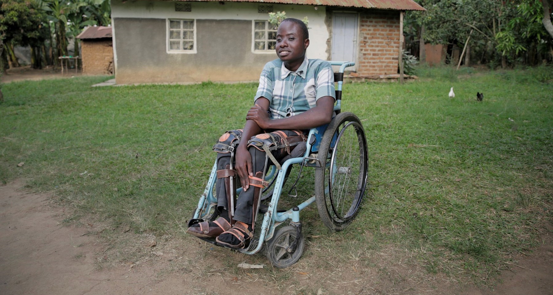 En ung, smilende gutt sitter i rullestol på gressplen med hus i bakgrunnen i Uganda. 