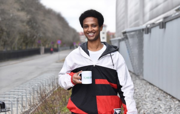 En ung mann står foran et grått bygg, og smiler til kamera. Han har en kopp hvor det står "Tøyen sportsklubb" i hånda.