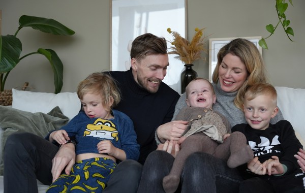 En familie med to voksne og tre barn smiler og sitter i en sofa.